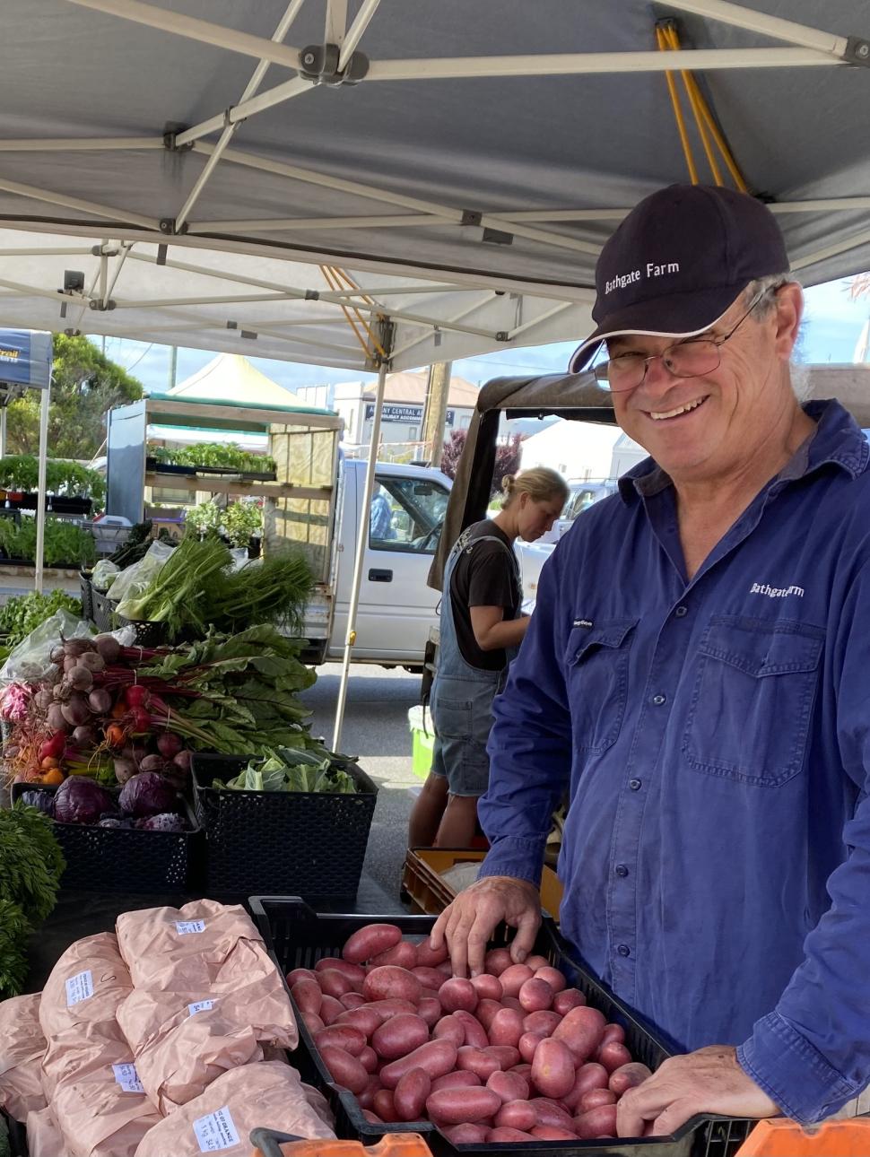 an adult man smiles at the camera surrounded by fresh produce at a stall