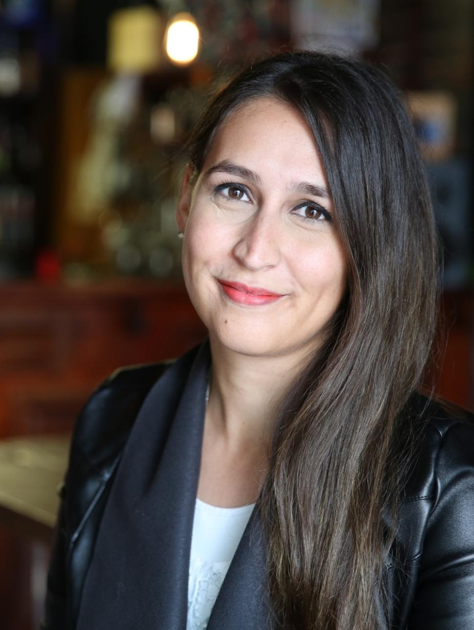 a person with long dark brown hair smiles at the camera wearing a white top and black jacket