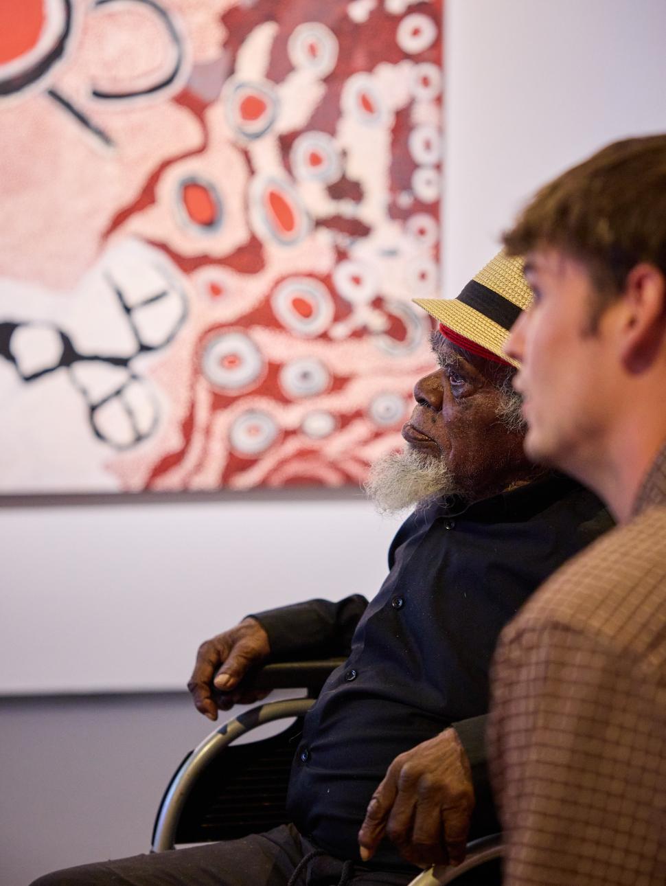 Old man in a wheelchair with an younger man next to him looking at a vibrant Aboriginal painting in an exhibition  