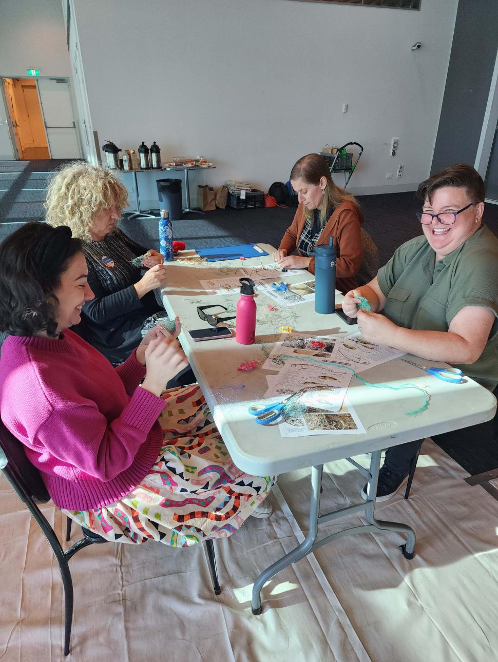 Four people sit around a fold-out table in a large white room with white tarpaulin on the floor. They smile as they craft small net sculptures.