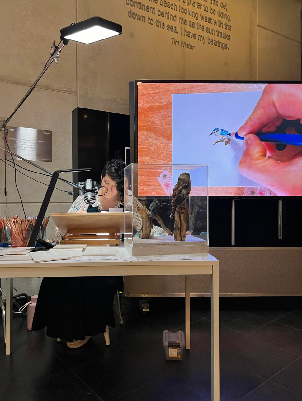 a person sitting at a desk drawing in detail with a screen behind showing the details 