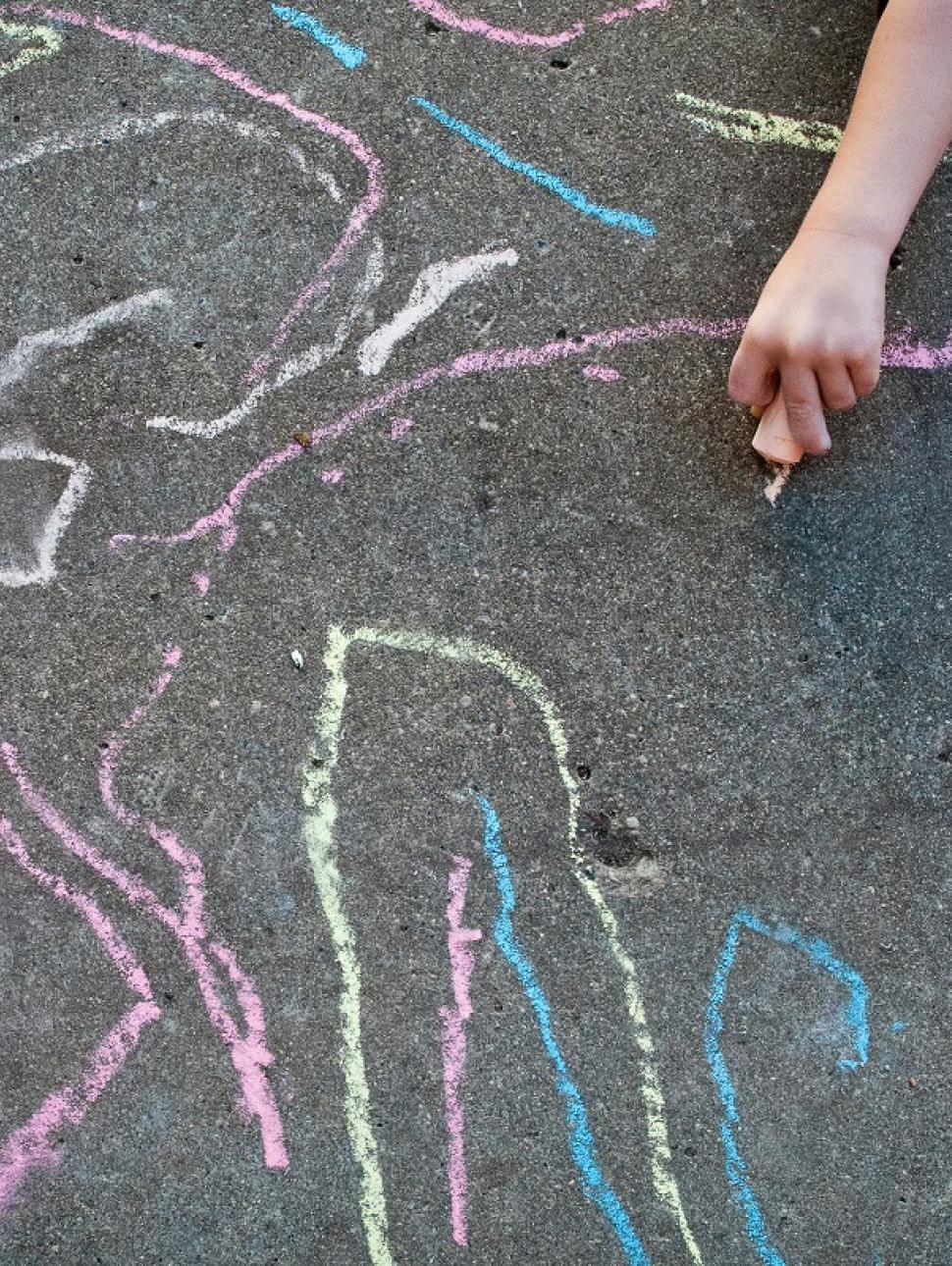 A child's hand holds an orange piece of chalk and draws on a road-like surface which is filled with colourful chalk lines