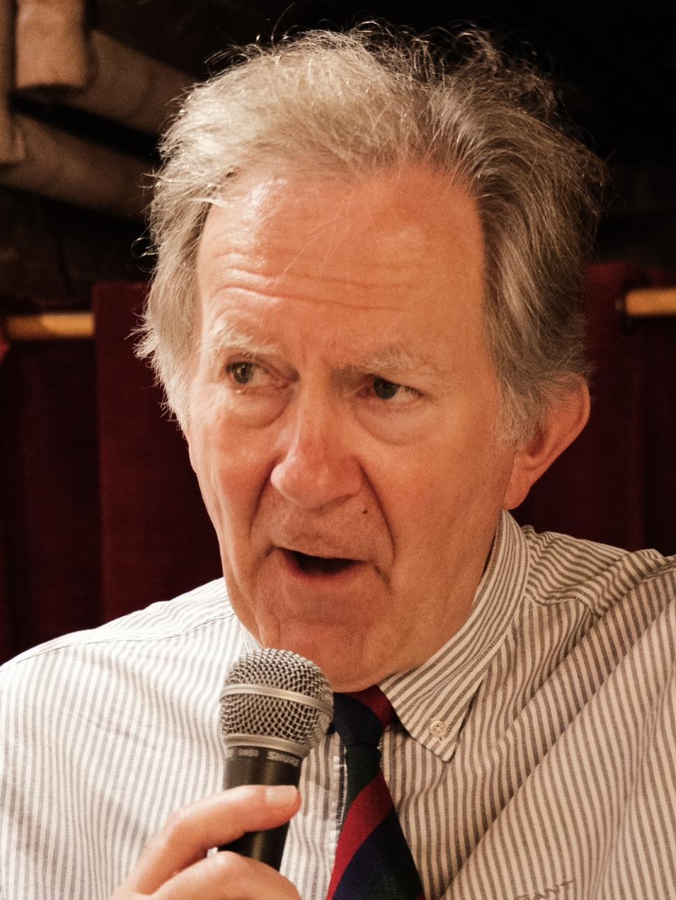 an adult wearing a white short and striped neck tie, holds a microphone and speaks with passion.