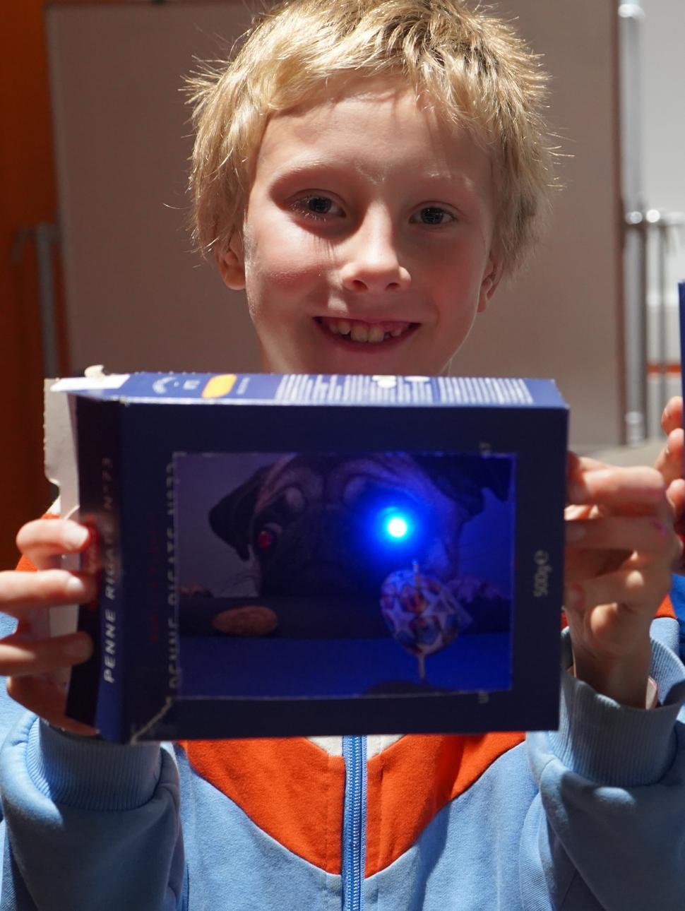 A young boy is holding up the theatre he made. It has blue LED lights in the display.