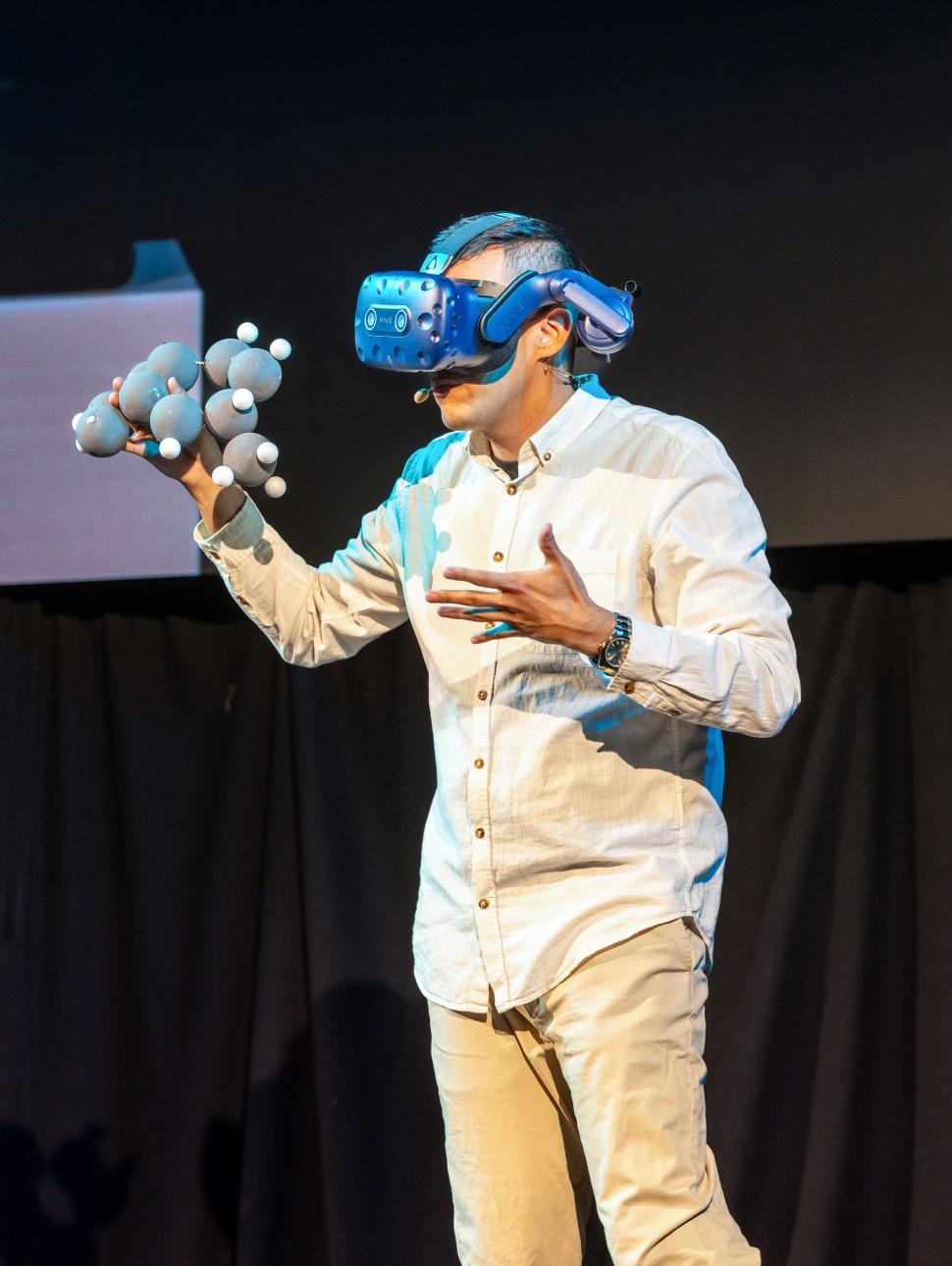 someone on stage wearing a VR headset, holding an atom diagram.