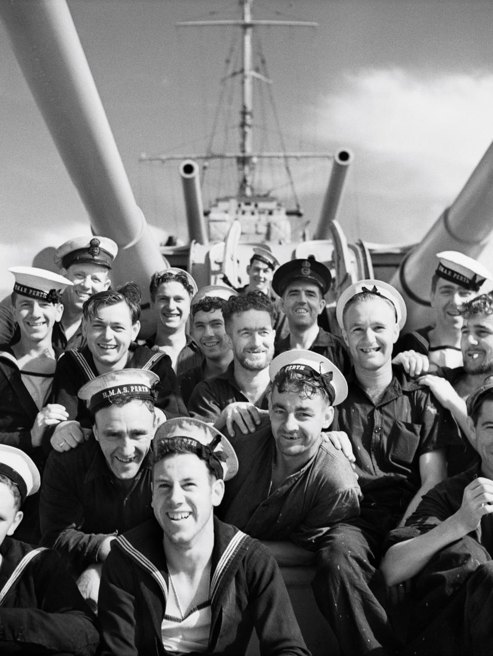 group of men in navy uniform on HMAS Perth smiling for a photo sitting under the ships guns