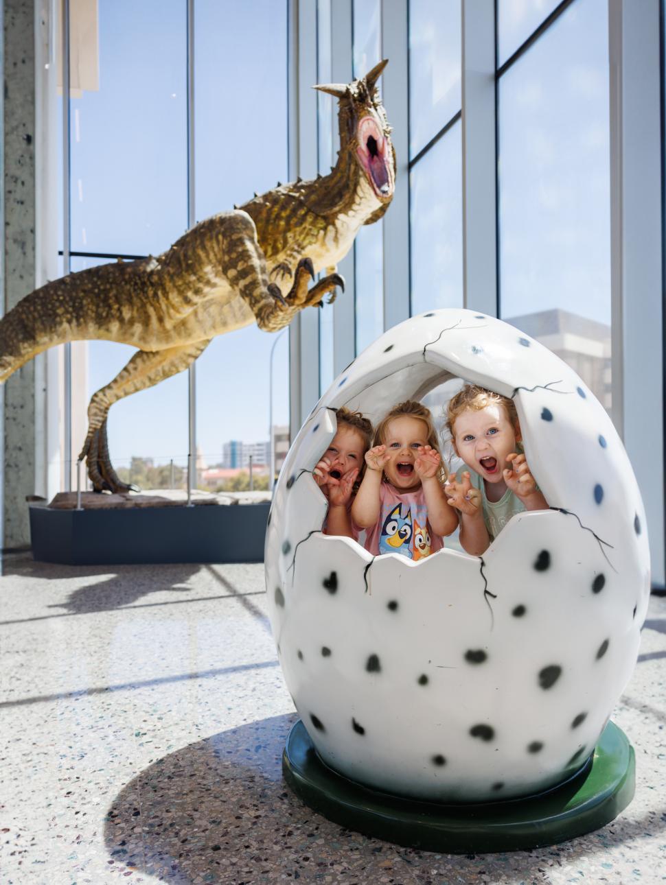 Three children inside a cracked dinosaur egg with a dinosaur model behind them in a museum.