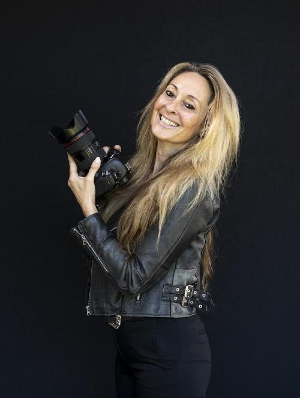 Photograph of Martine Perret, who has long blonde hair and wears a black leather jacket, posting in front of a black background with her camera in hand