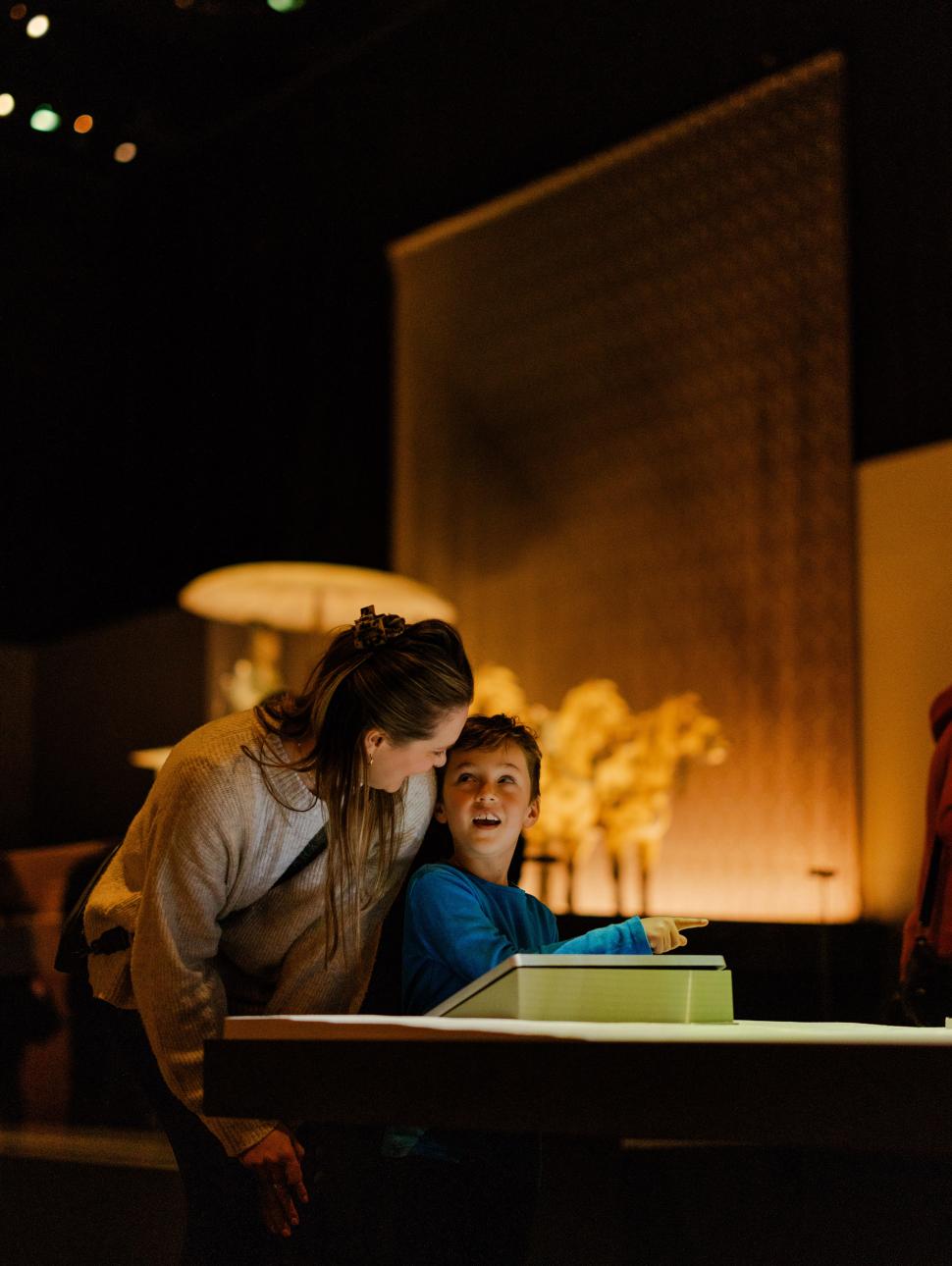 An adult and a child looking at an illuminated display table in a dimly lit room with warm lighting