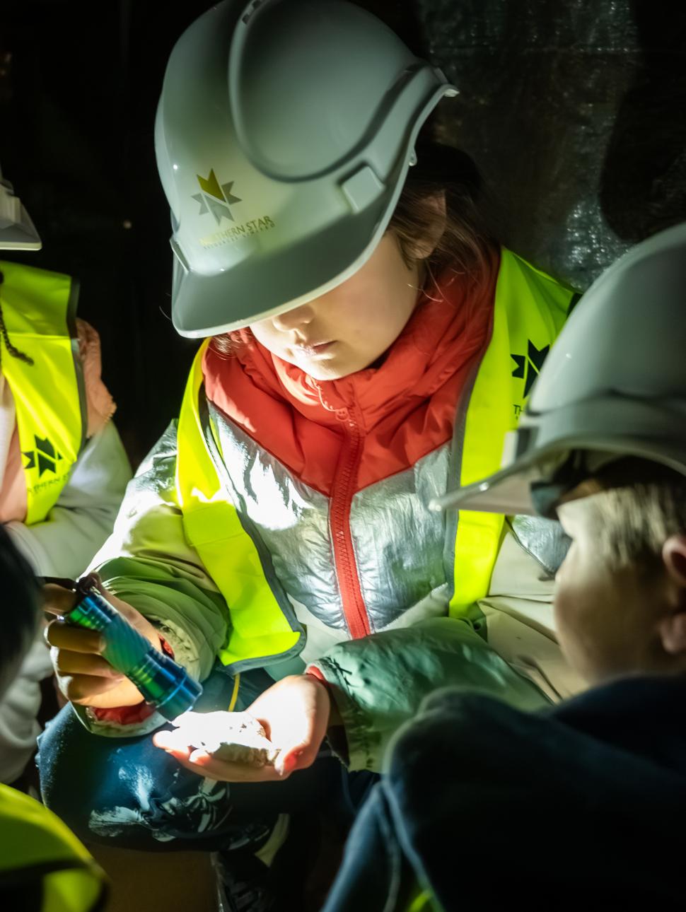 Children wearing hard hats and safety vests, examining an object in dim light.