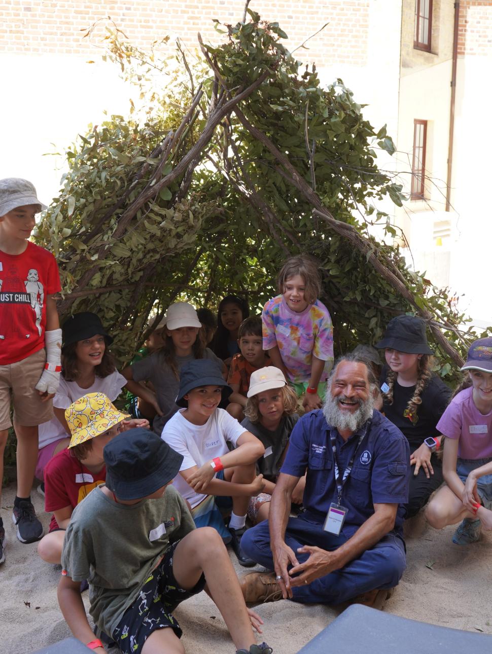 Derek Nannup and a group of kids sitting underneath a kalak (shelter)