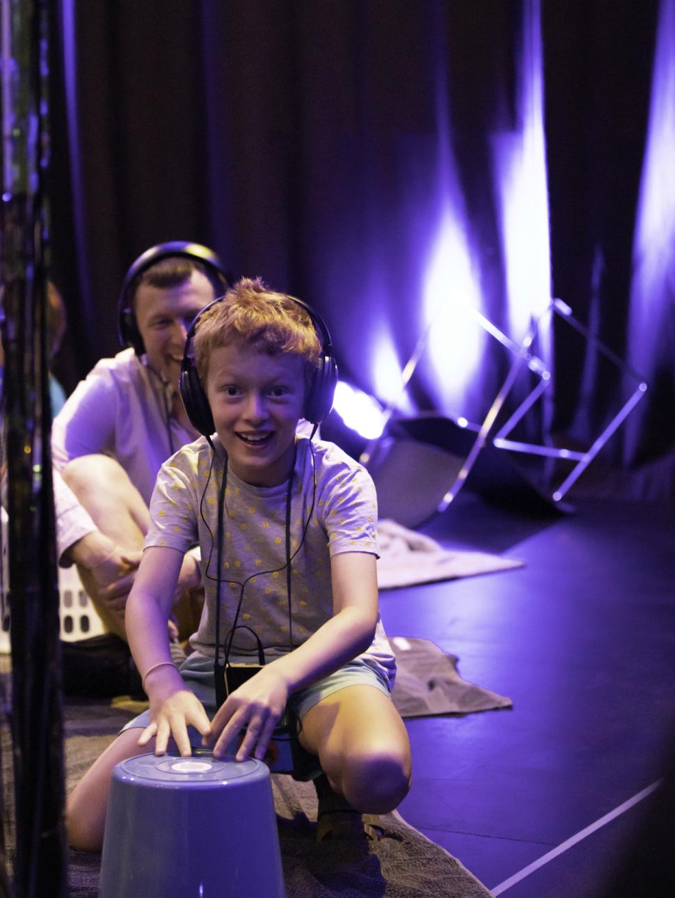 Smiling child wearing headphones squatting on blanket with upturned bucket in front of them. Smiling adult behind them also wearing headphones.