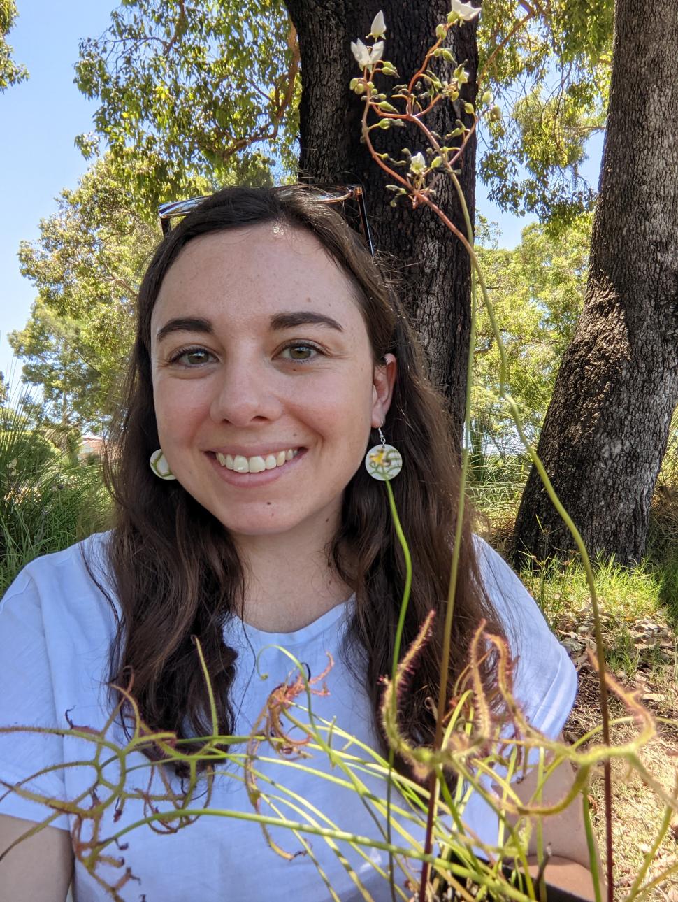 Image shows a close up of Laura Skates the presneter for this Meet the Museum session. She has long dark hair ans wearing a grey T-shirt. Bushland is in the background 