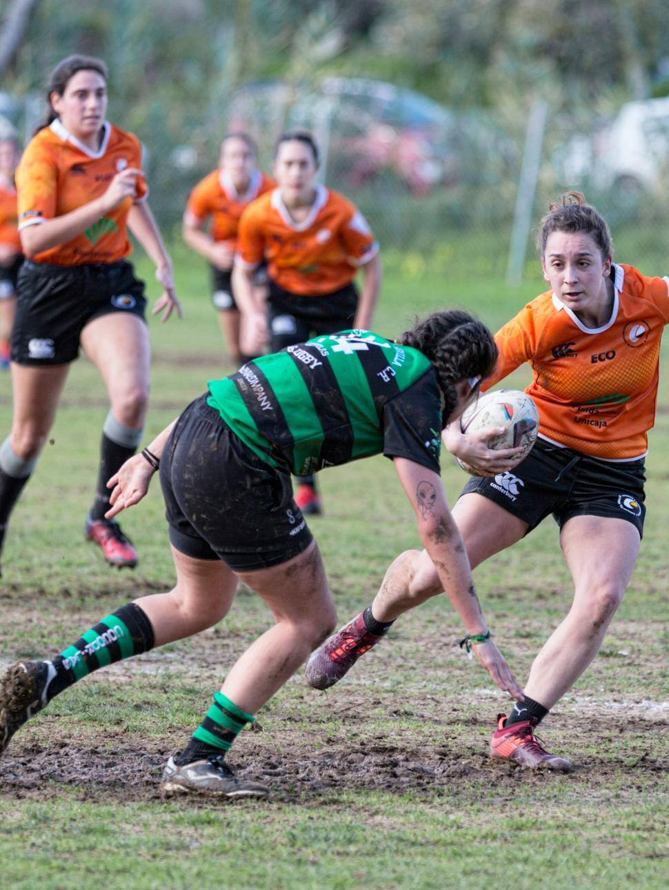 Image shows two competing teams of women playing rugby. ONe team has an orange uniform and the other a green and black striped uniform. The playing field is muddy.