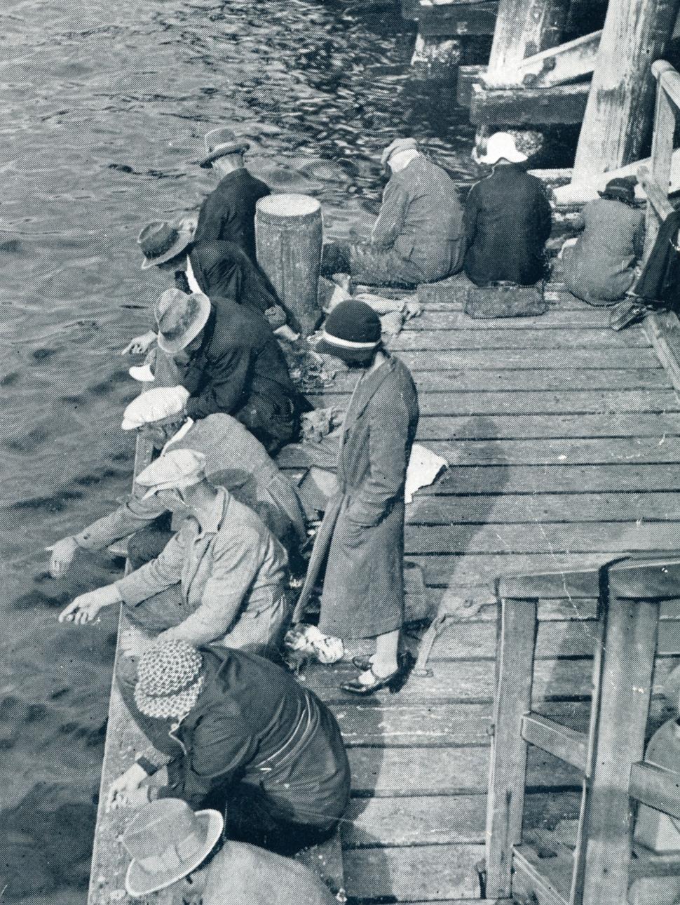 An archival photograph showing a group of people are leaning over a dock and pointing in the water 