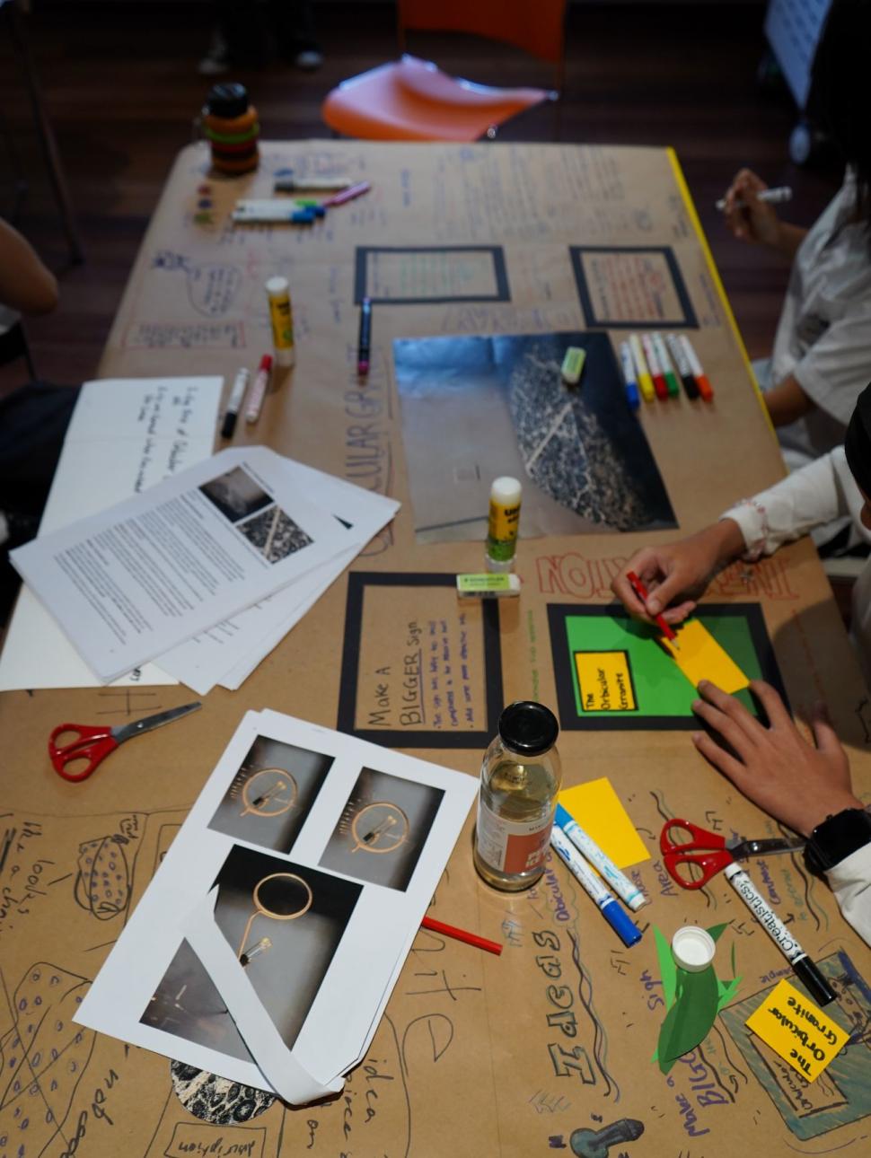 The image shows a long table covered in brown paper and various materials that students are using for a design activity.