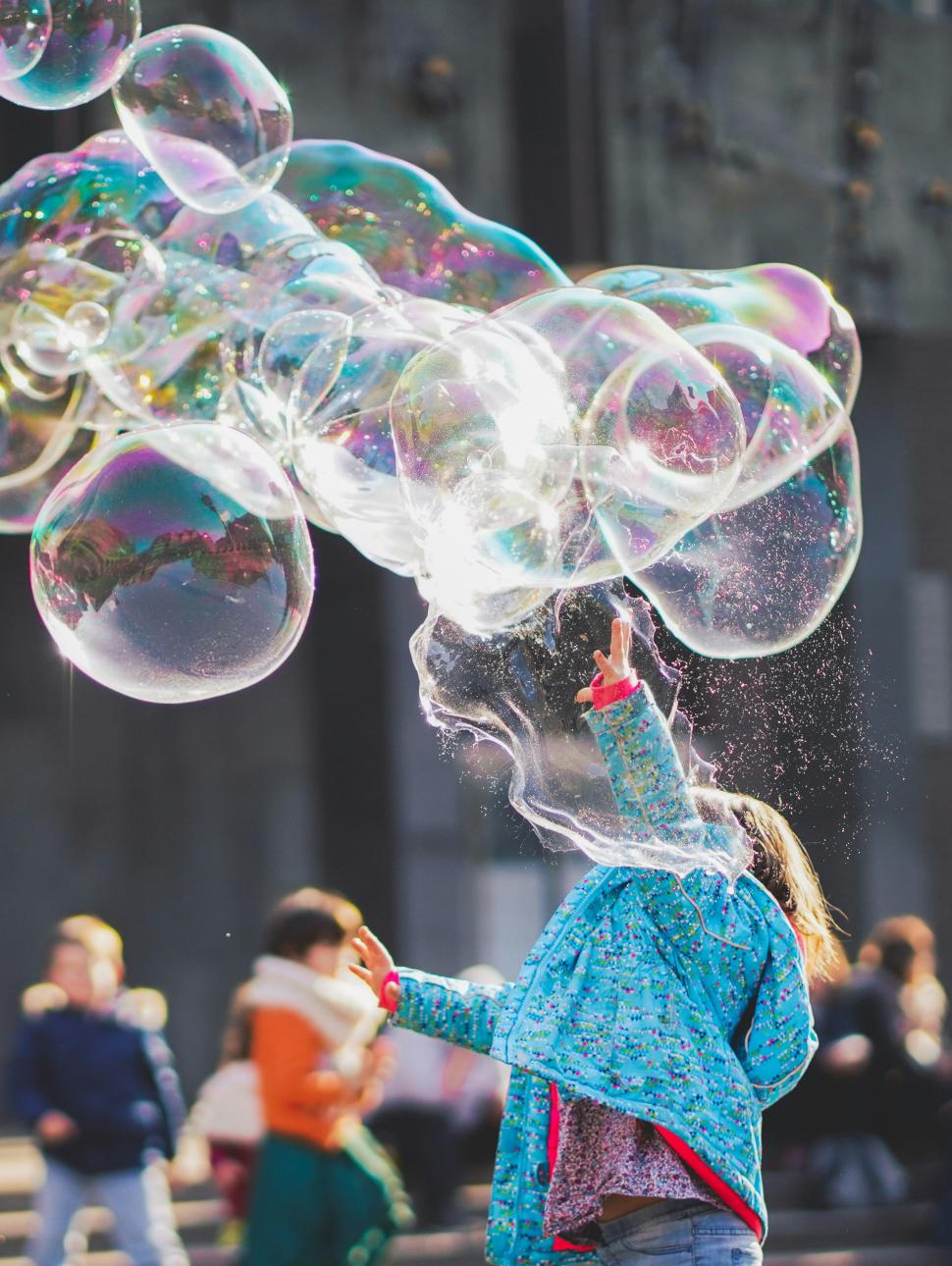 Child in blue jacket reaches into the air to touch giant bubbles floating past