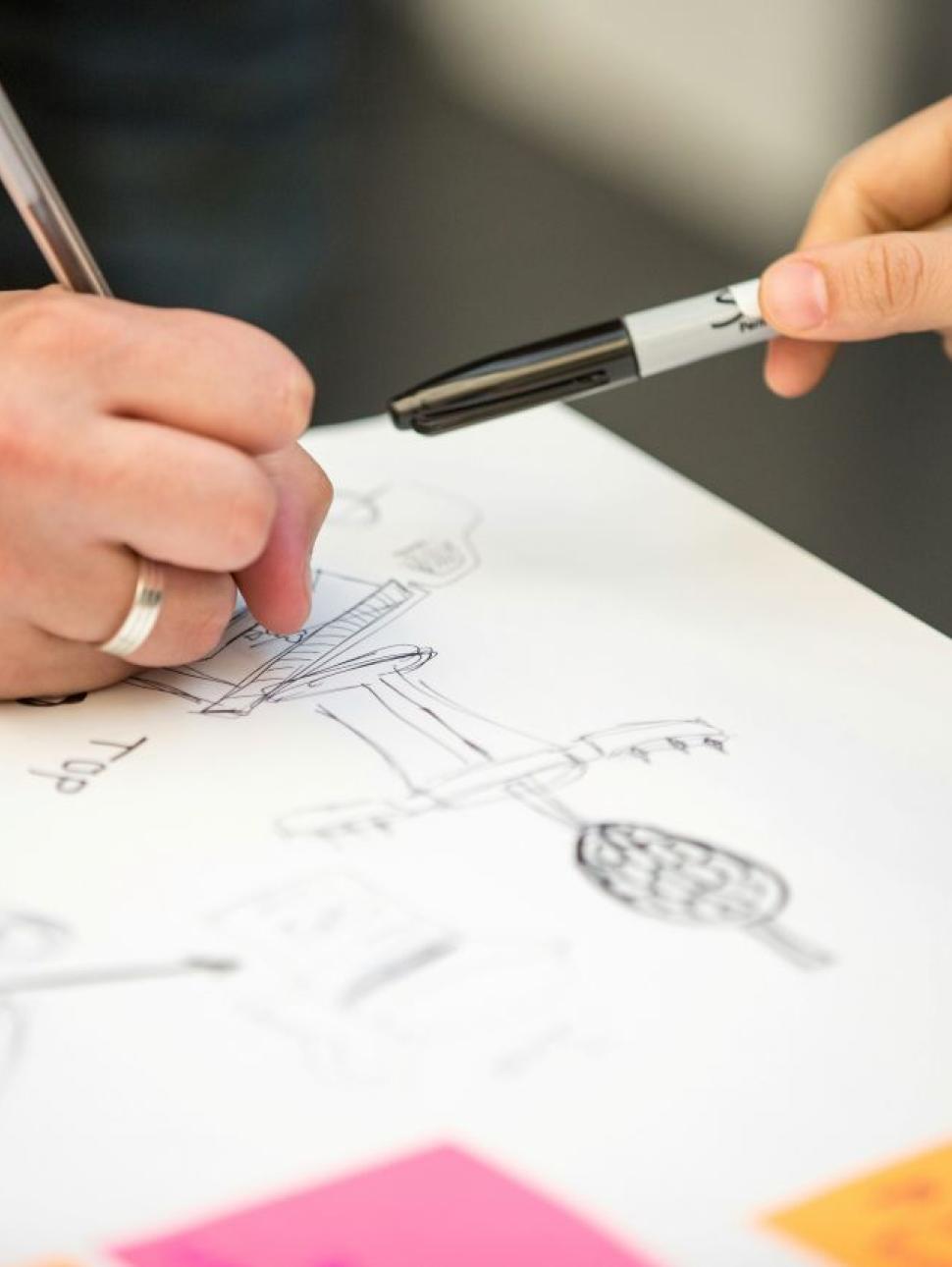 Image shows a close up of peoples hands as they sketch out their ideas on paper.