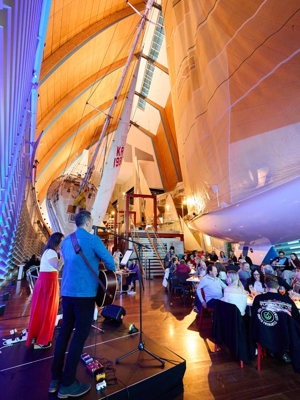 people seated on tables, performers on a small stage in the foreground with yachts in the background