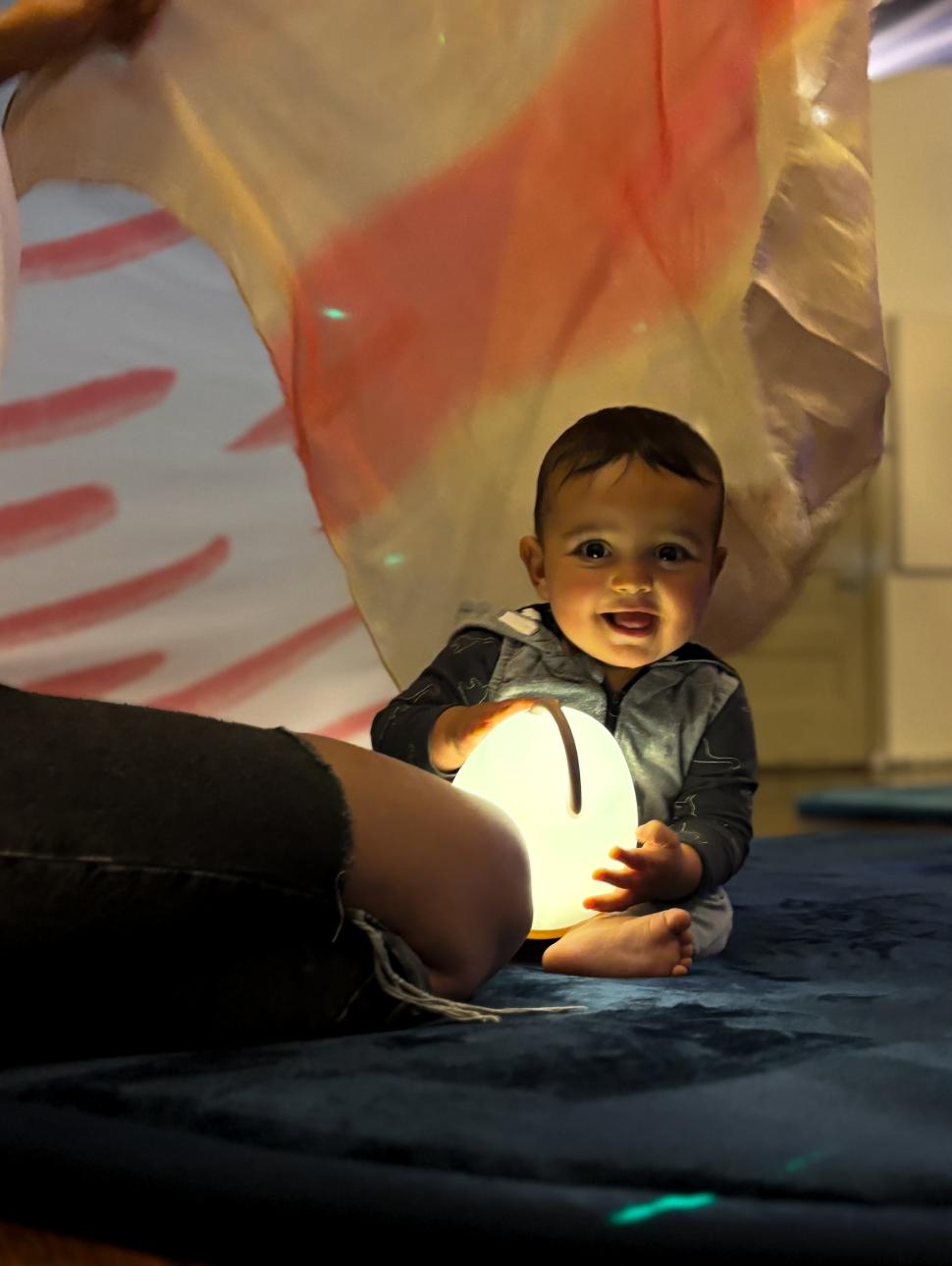 Smiling baby sitting on rugwith egg-shaped lamp. Adult kneeling next to them