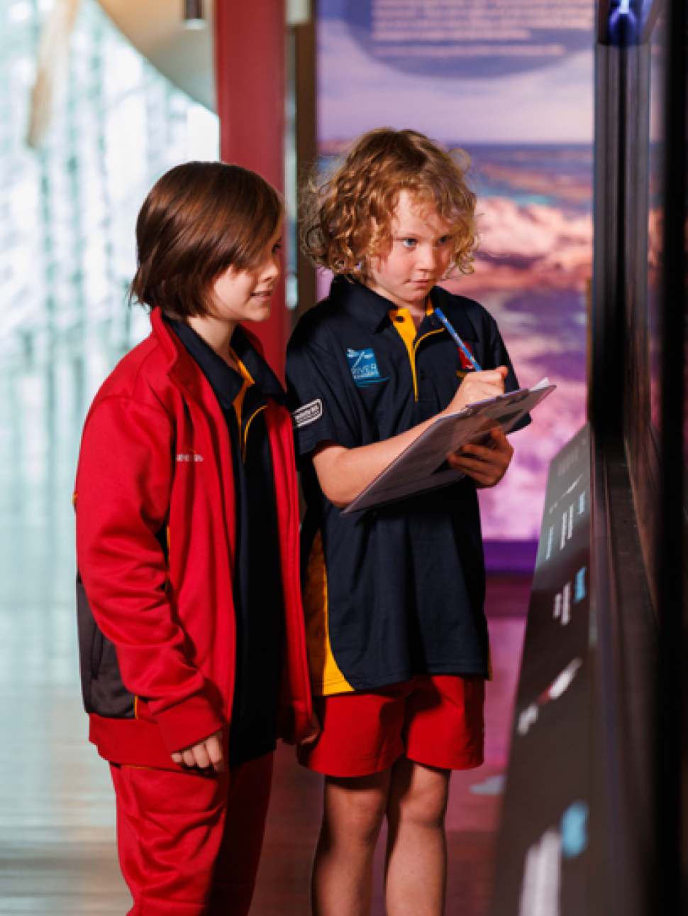 Two students in school uniforms are standing in front of an exhibit display, actively participating in an educational activity. 