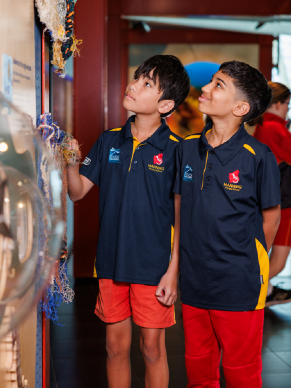 Two students in navy and yellow school uniforms are closely examining a textured display featuring woven materials and fibers. The exhibit appears to showcase traditional or natural elements, possibly related to cultural heritage or environmental themes. The setting is indoors, with bright lighting and other students visible in the background, suggesting an educational excursion or museum visit focused on interactive learning.
