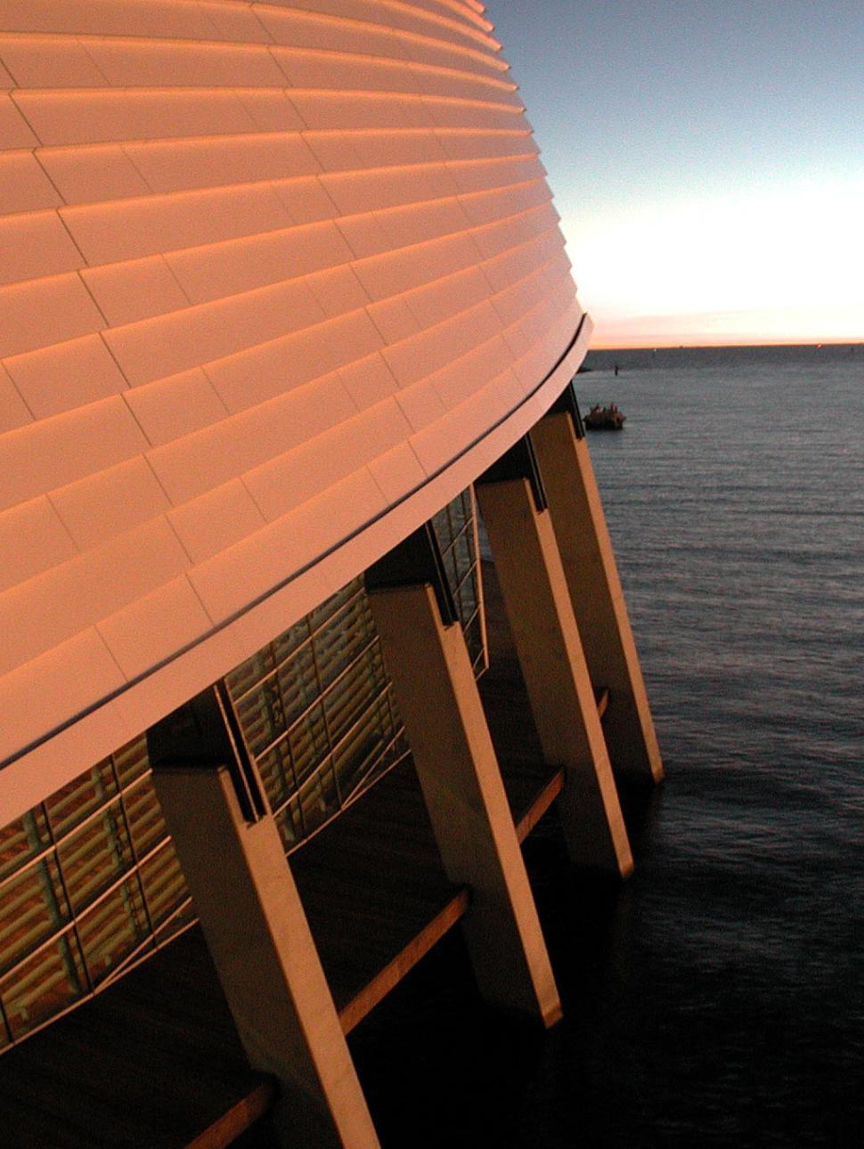 Half the photo pictures the outside of the museum - its curved roof, windows, boardwalk and supporting concrete beams. The other half pictures a river. There a few clouds in the distance that look orange in colour as the sunsets.