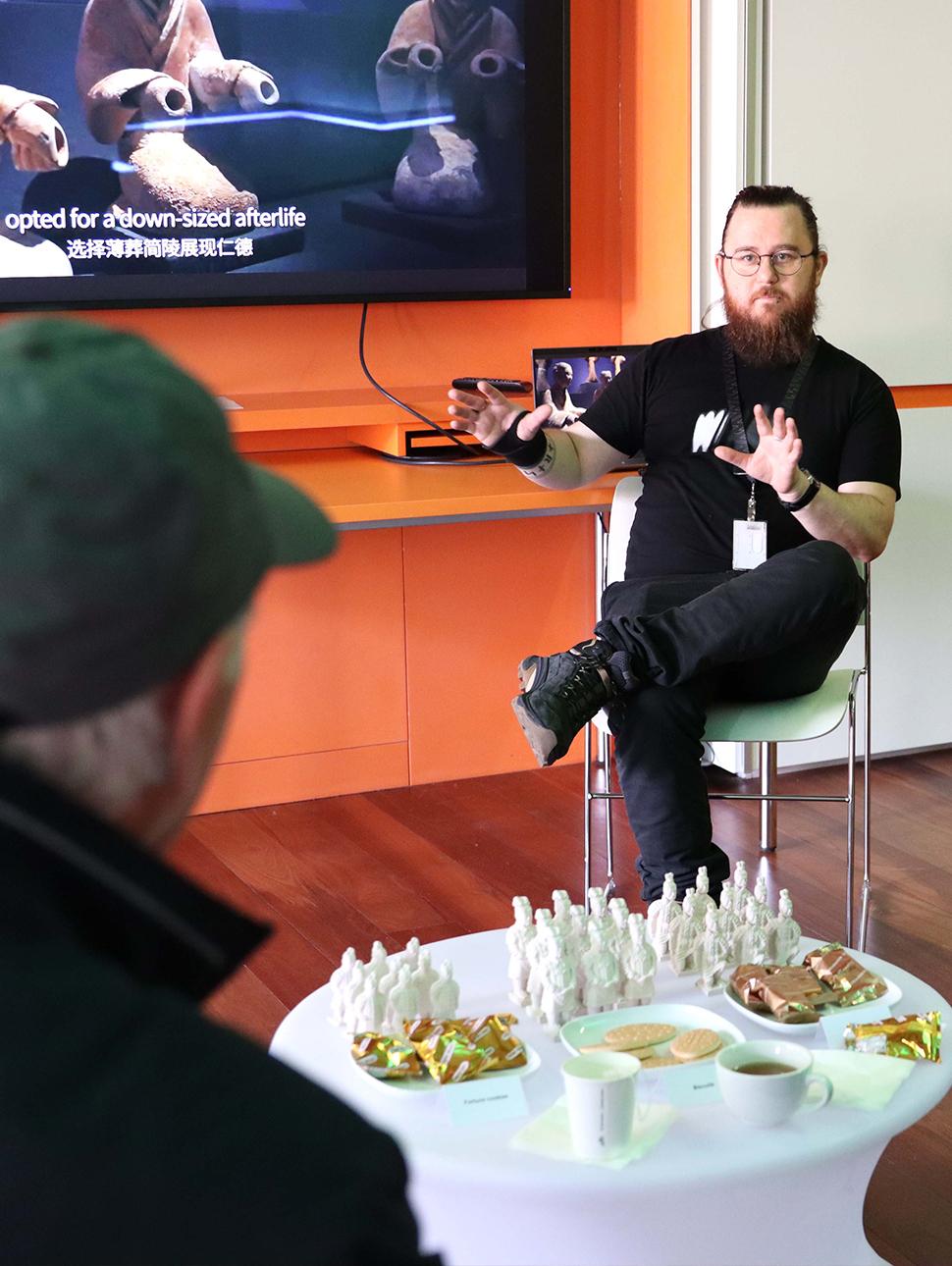 A man with a beard sits on a chair presenting information and gesturing, there is a coffee table with artefacts and tea and biscuits on it in the foreground.