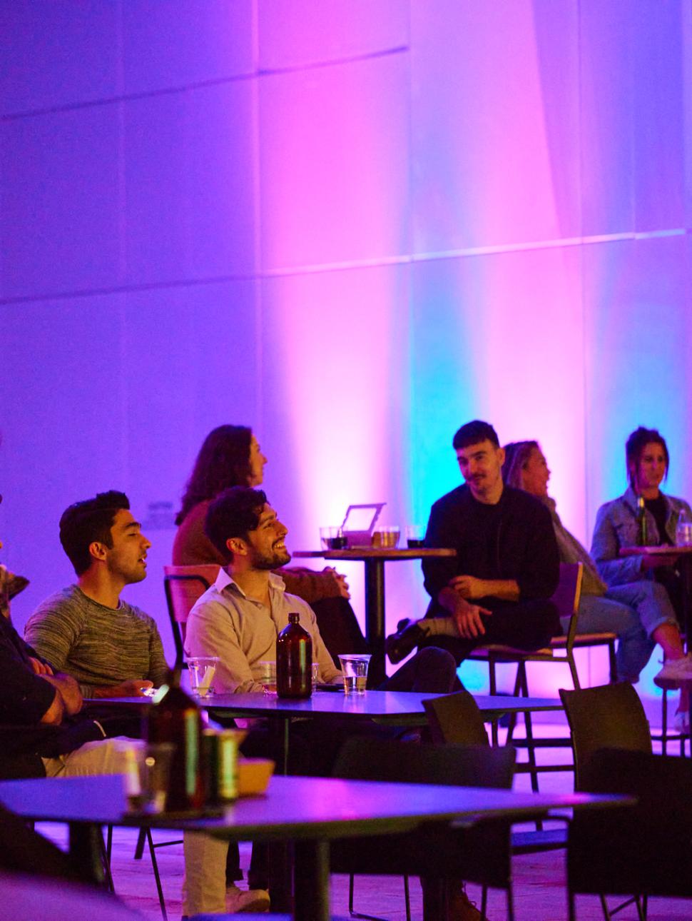 People seated at tables in a dimly lit room with purple and blue lighting, engaged in conversation at a social event.