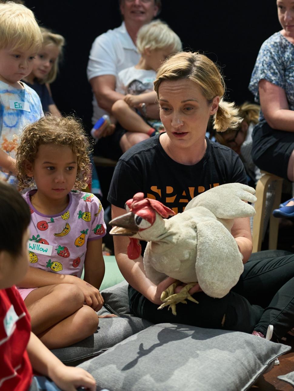 a group of children sit around an adult with a puppet 