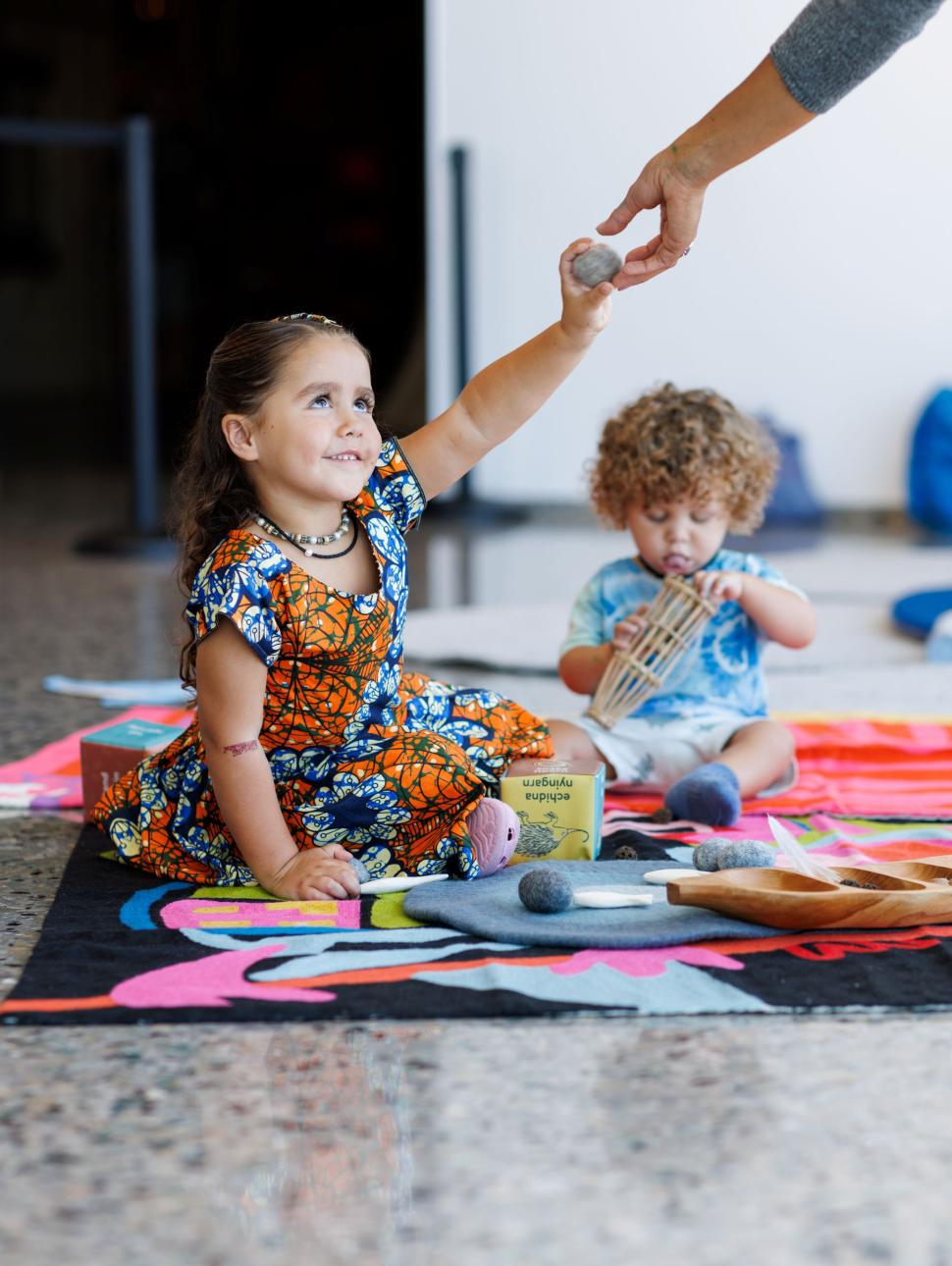Two young children sitting on a mat - someone passing something to the young girl.
