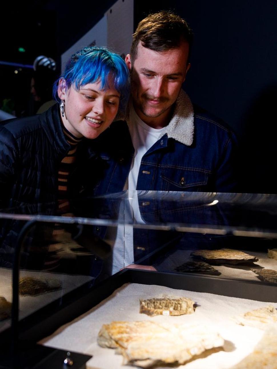 A woman with short blue hair and a man with short brown hair and a moustache smile as they lean over a glass case of objects.