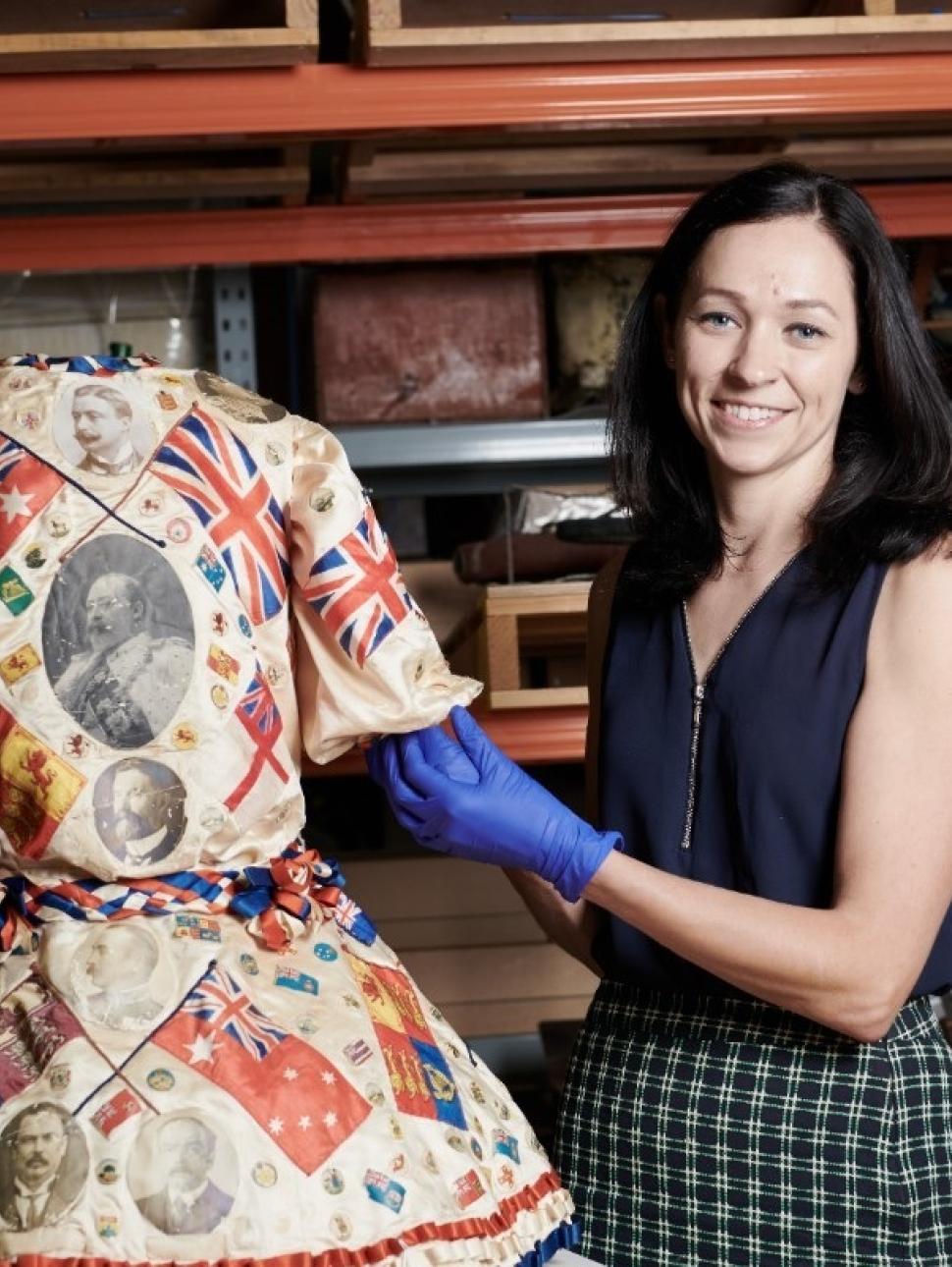 a person with archive gloves stands next to a colourful dress garment on a mannequin 