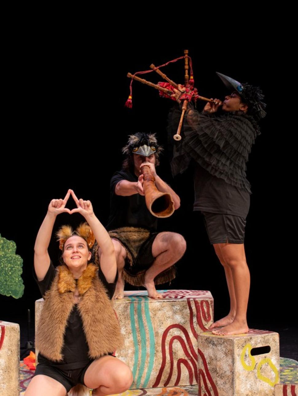 Yirra Yaakin Theatre Company present Djinda Kaatijin. This production image shows a trio of young Aboriginal theatre performers, one seated with her hands in a star formation above her head; one crouched nearby on a stage block playing didgeridoo, and the third performer standing on a stage block playing the bagpipes.