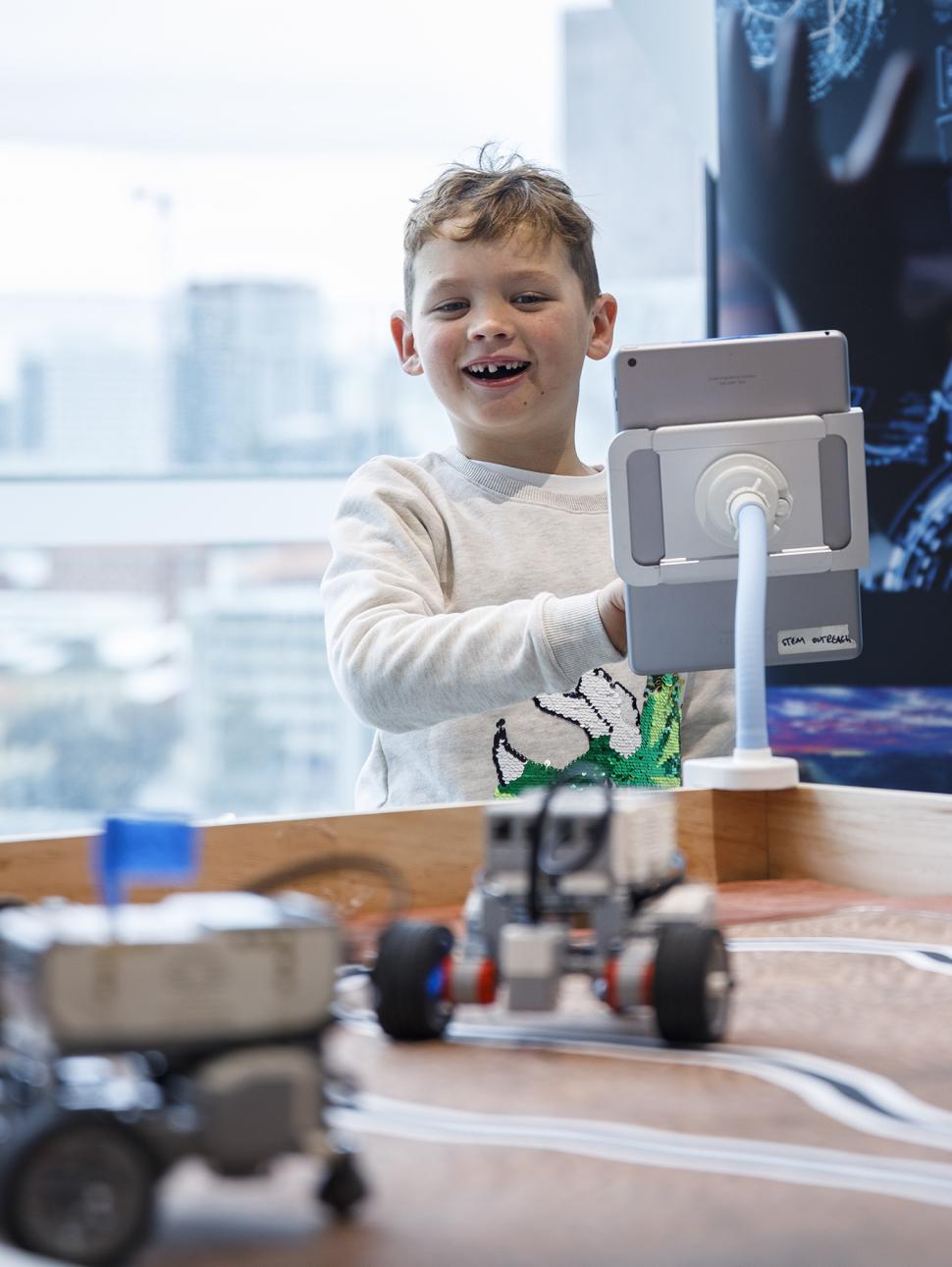 Young child standing in front of an iPad smiling. Robots on table in foreground