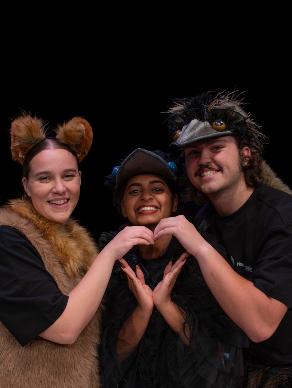 Three performers from Yirra Yaakin' 2026 production of Djinda Kaatijin, shown together in costume (Emu, Crow and Dingo) all smiling and making a loveheart shape with their six hands together.