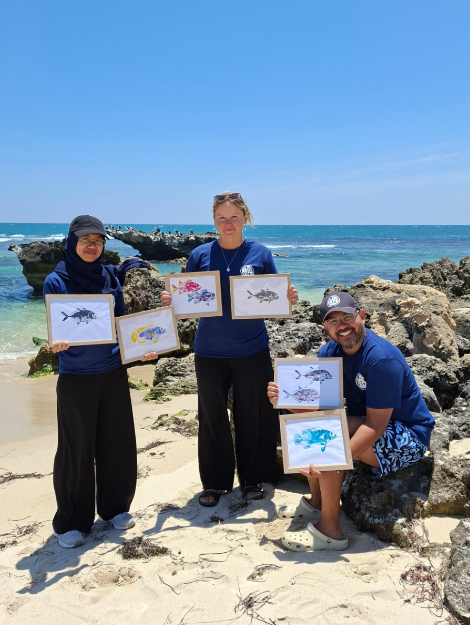 three people are on a beach with the ocean behind them. One person is sitting on a rock, the others are standing' All art holding some artwork showing fish. 