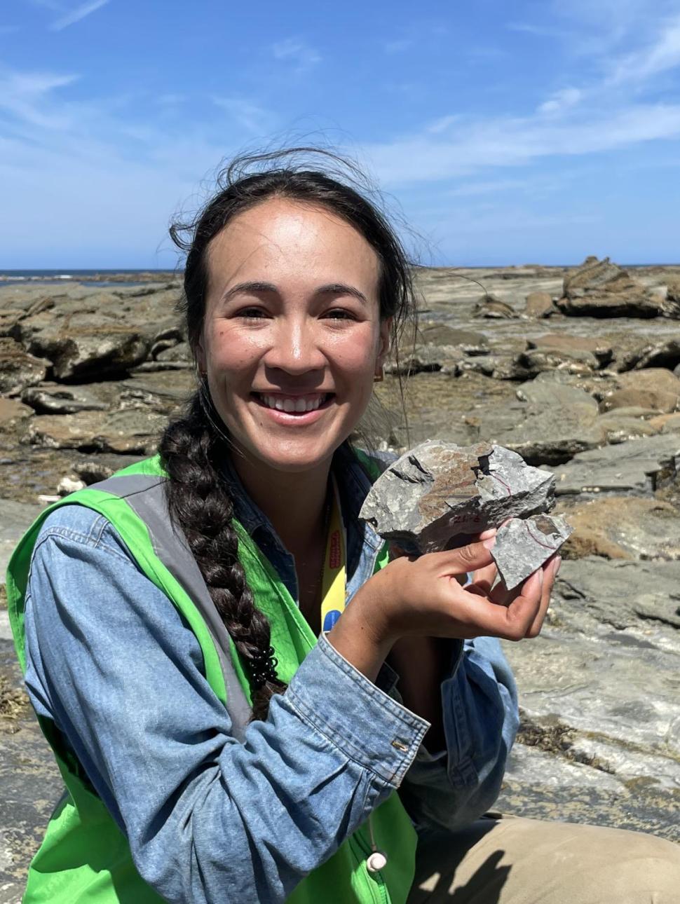 an adult person outside in a rocky landscape holds up a rock with some fossils inside the rock