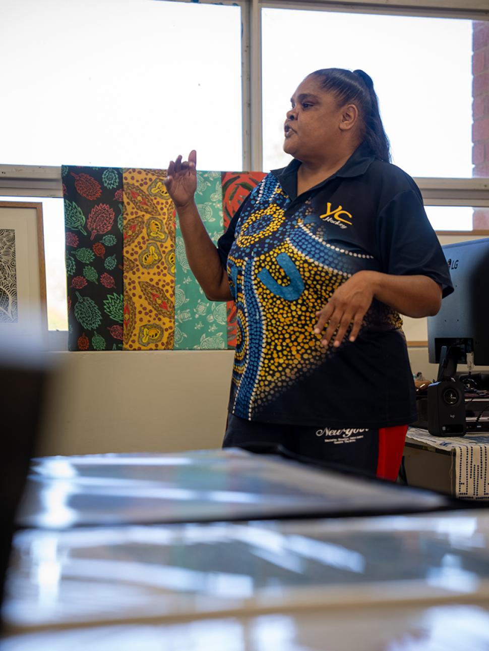 a person stands and is talking facing the side and in the background artworks are being displayed