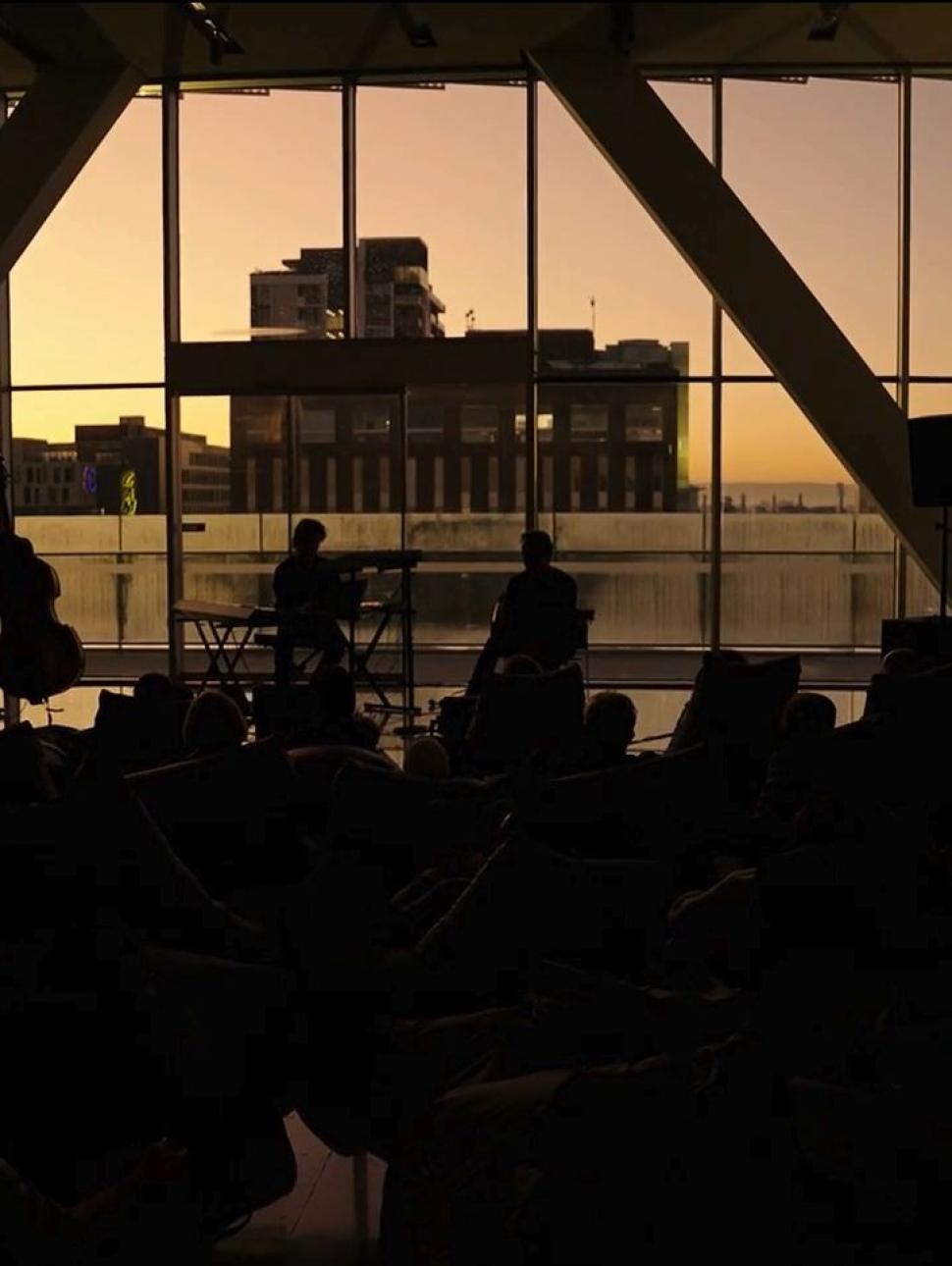 Silhouetted musicians perform for a seated audience inside a large glass-walled venue at sunset, with city buildings visible through the windows.