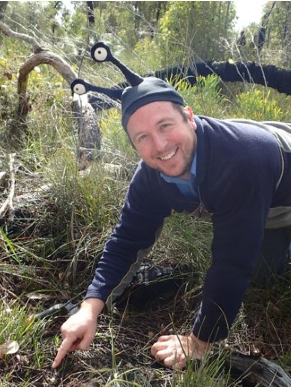 a person out in the field pointing to the ground where they have located a snail. The person smiles and is wearing a snail hat