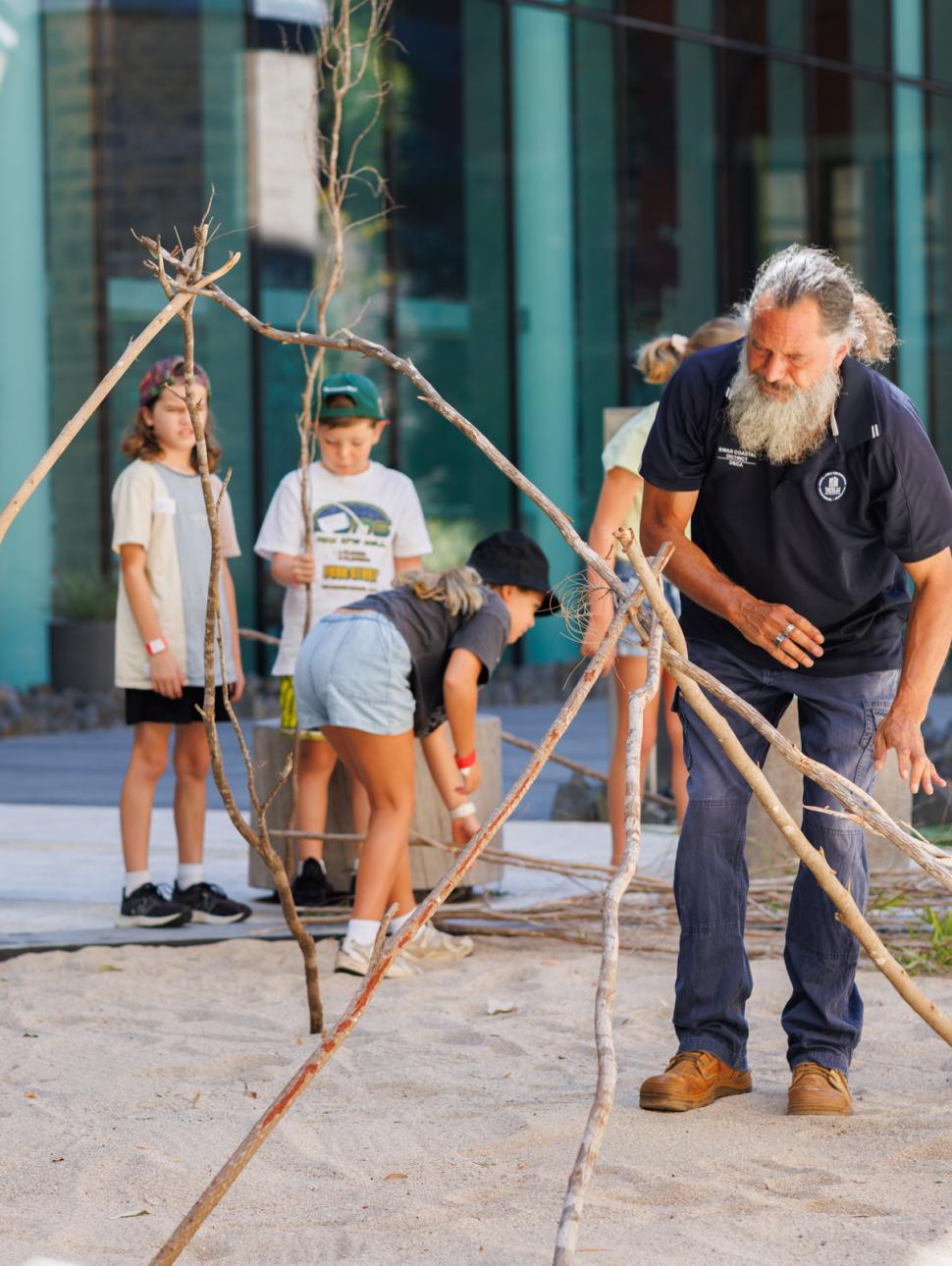 Man building a stick shelter while kids watch