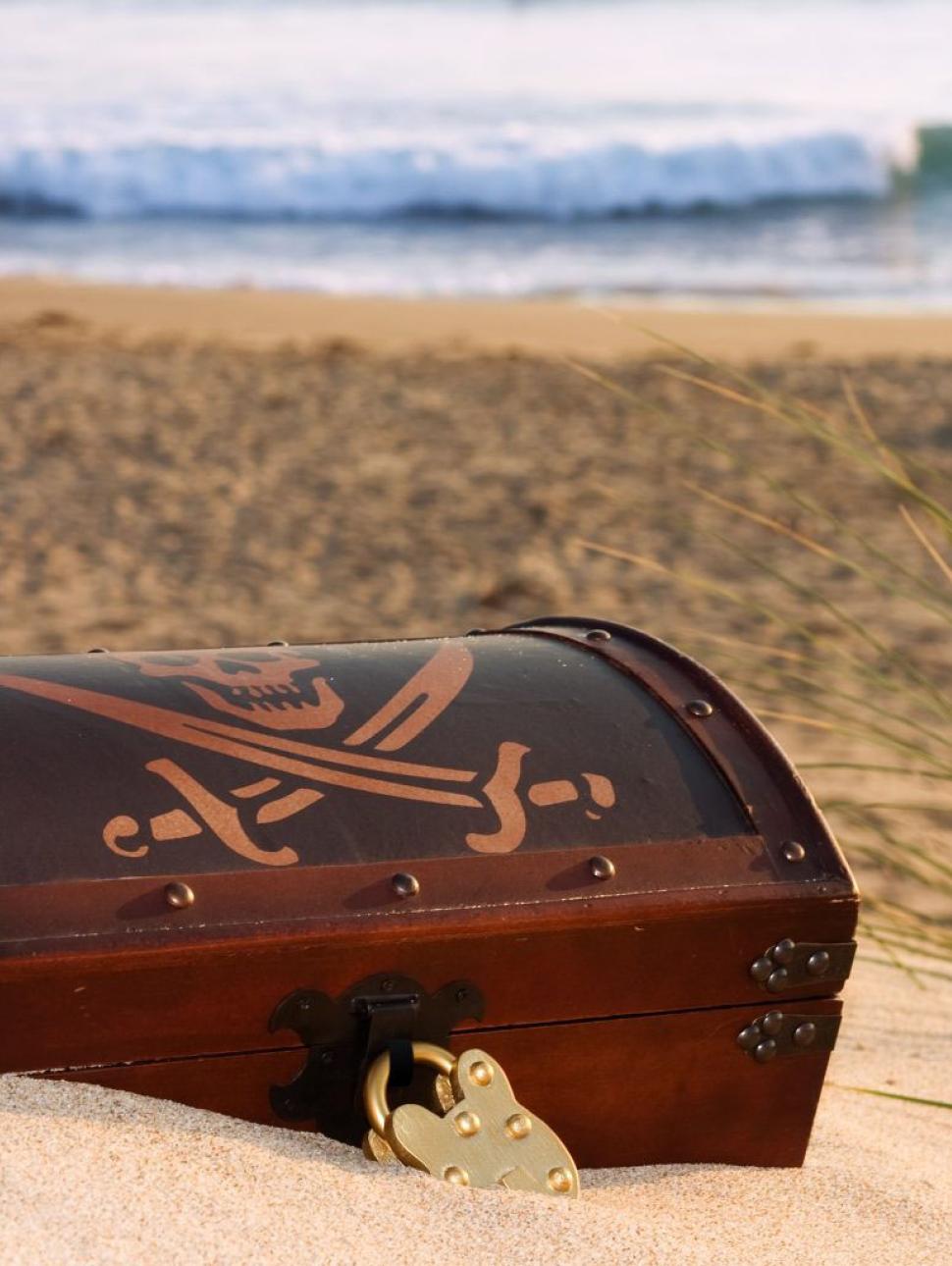 A treasure chest buried in sand on the beach with crossed swords and a skull showing on the lid, linked to the Treasures of the Deep event for Totally Lit 2026 festival.