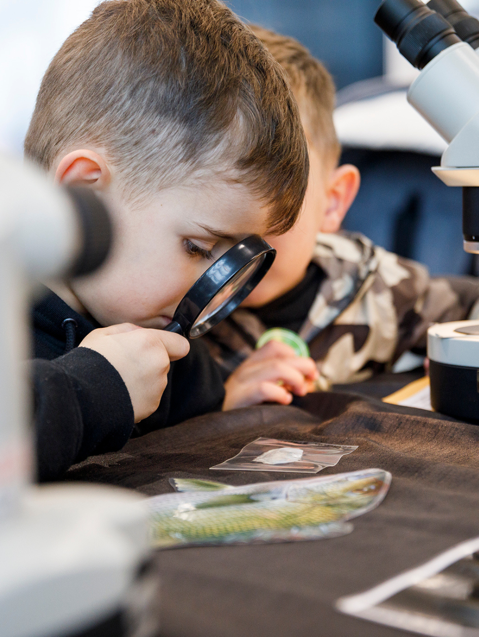Kid looking through magnifying glass at object with telescope behind him