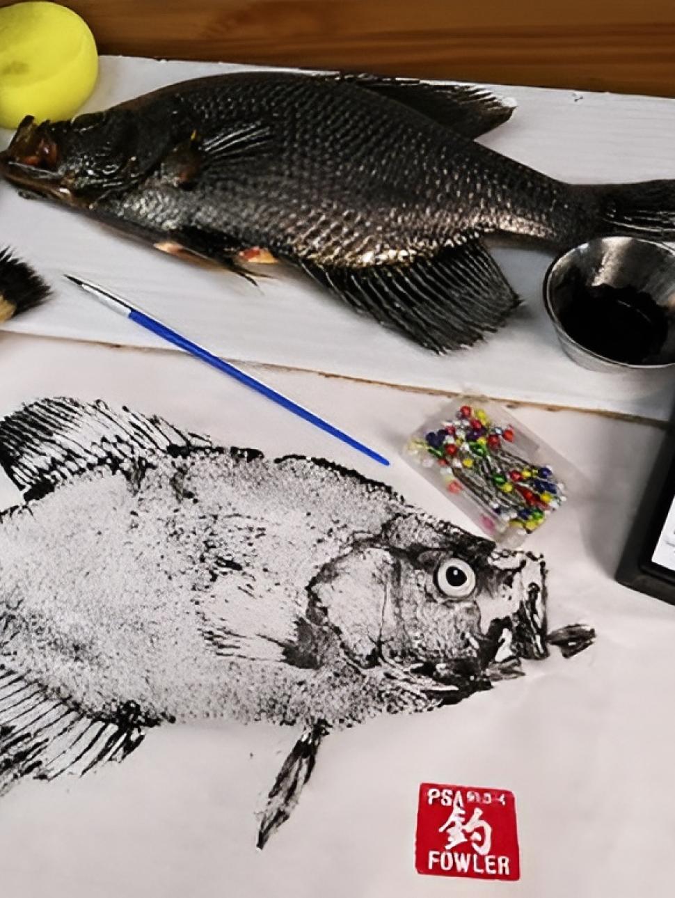 a top view of a table with a fish print and a fish with materials used to create the artwork