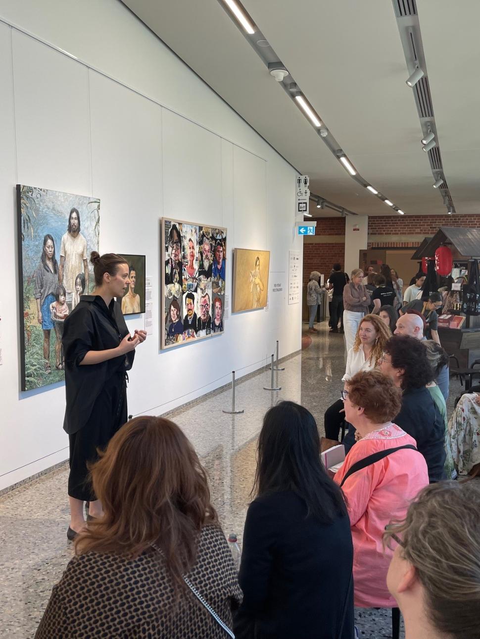 A woman with a black shirt and brown hair pulled into a bun is standing in front of a wall covered in paintings. She is speaking to a group of people sitting on stools in front of her. 