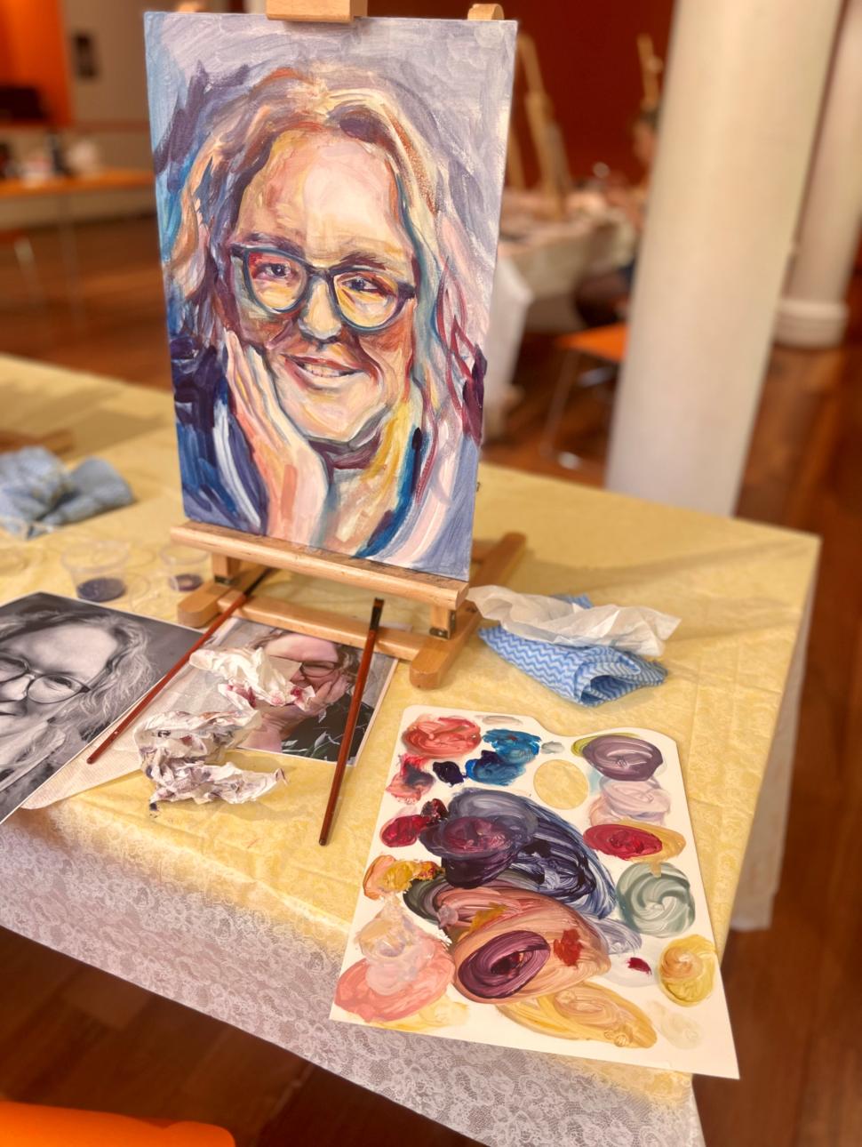 A painting of a woman's face sits on an easel on the table. The woman has grey hair and a blue shirt on, she is smiling and propping up her chin with her hand. Around the painting on the table is a pallette filled with paints and a selection of brushes 