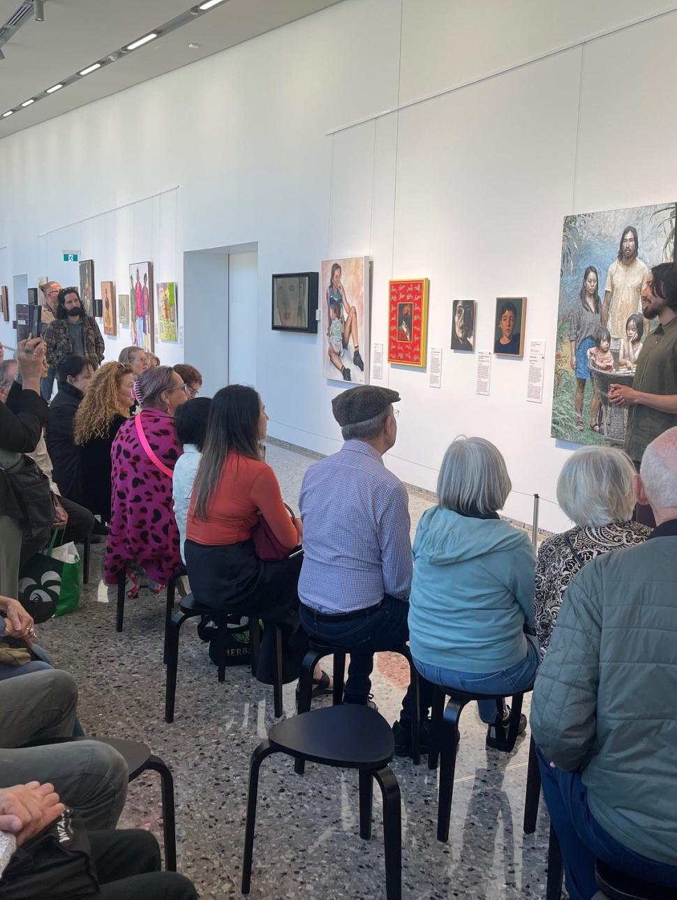 a group of people sit on black stool facing away from the camera and towards a selection of portraits on the wall. they are listening to a young man with black hair and a beard standing next to a large paitning. 