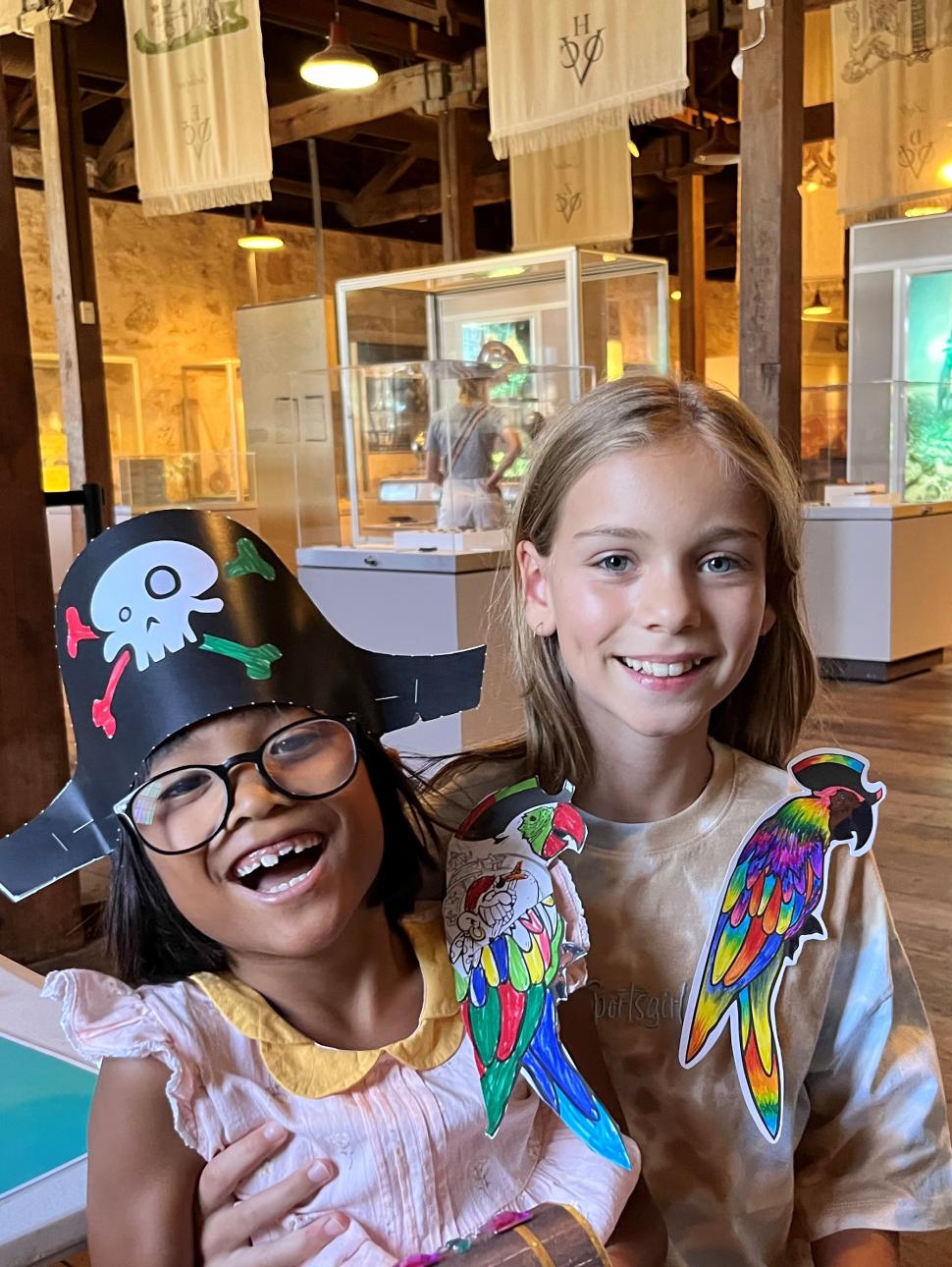 Two girls stand together smiling, wearing paper parrots on their shoulders. One is wearing a pirate hat too.  Behind them are display cases, a lightbox with a diver and historical flags are hung from the roof. The floor is wooden and the walls made of limestone. 