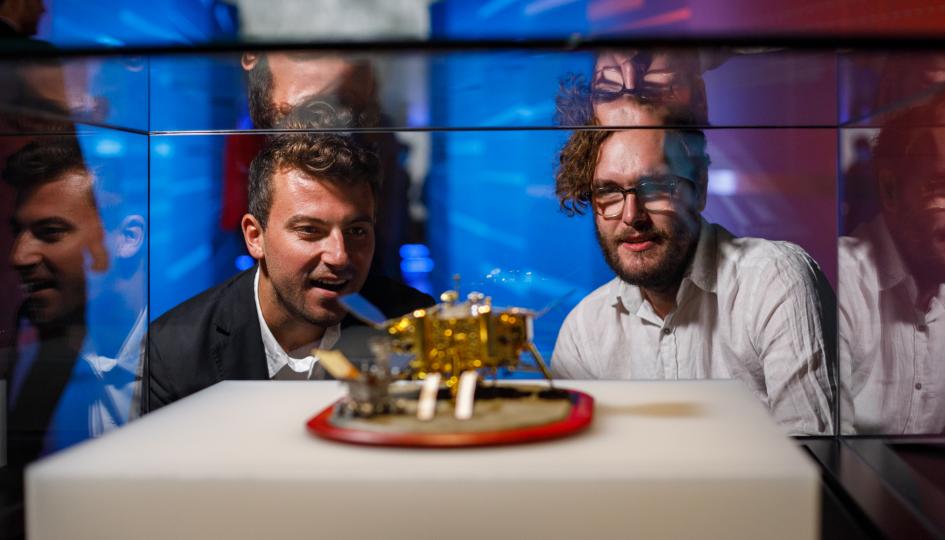 A person wearing a black suit jacket with short-cropped curly hair and a person with a white button up shirt, short hair and thin rimmed glasses stare with expressions of interest at a small gold spacecraft model in a glass Museum showcase